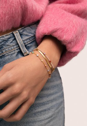 Muñeca con pulseras doradas y de cuentas apiladas, con vaqueros azules y manga de suéter rosa peludo.