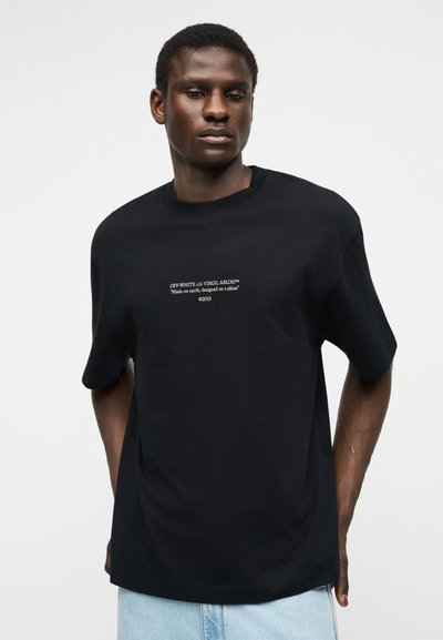 Νέος άνδρας που φοράει μαύρο μπλουζάκι Off-White με λευκό κείμενο στο κέντρο, συνδυασμένο με ανοιχτόχρωμα τζιν, στέκεται μπροστά σε απλό φόντο.