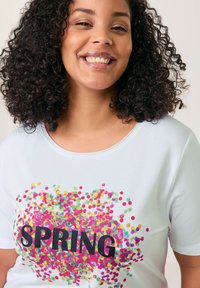 Weißes Baumwoll-T-Shirt mit bunten Punktmustern in Rosa, Grün und Gelb um das Wort "FRÜHLING" in fetten schwarzen Buchstaben.