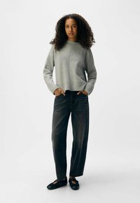 Grå stickad tröja med hög hals, i kombination med mörka, löst sittande jeans och svarta platta skor. Slät textur, avslappnad design.