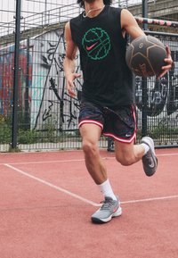 Joueur de basket en débardeur noir avec un logo pixellisé, short coloré à motifs, et chaussures Nike grises, dribblant un ballon de basket.