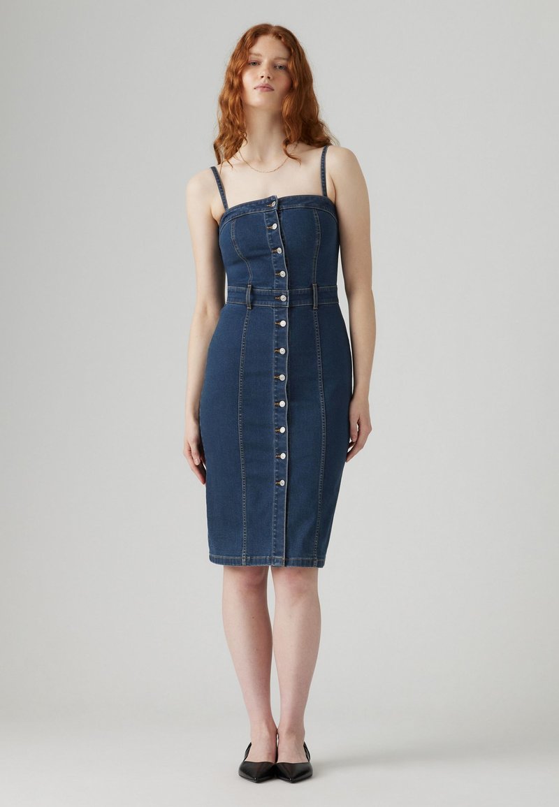 Vestido de mezclilla en azul oscuro con forma ajustada, tirantes finos, botones en el frente y cintura ceñida. Textura suave y diseño a la altura de la rodilla.