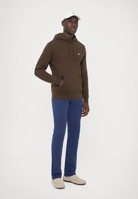 Ruda bliuzonas su priekinė kišene ir raišteliu, derinamas su mėlynomis kelnėmis ir smėlio spalvos batais. Balta kepurė su logotipu. Švarus, minimalistinis dizainas.