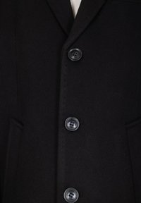 Indumento esterno nero con un tessuto testurizzato, caratterizzato da un colletto a revers, due tasche frontali e quattro bottoni neri al centro.
