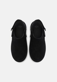 Sorte semskede slip-on sko med avrundet design, som har en glatt tekstur og diskret logo på innersålen. Ingen synlig maskinvare eller detaljer.