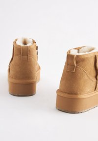 Paire de bottines en daim couleur camel avec semelles épaisses en caoutchouc et doublure blanche duveteuse, vues de dos sur fond blanc.