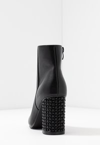 Czarny skórzany botek za kostkę na wysokim słupku z okrągłymi wstawkami, pokazany z tyłu na białej powierzchni.