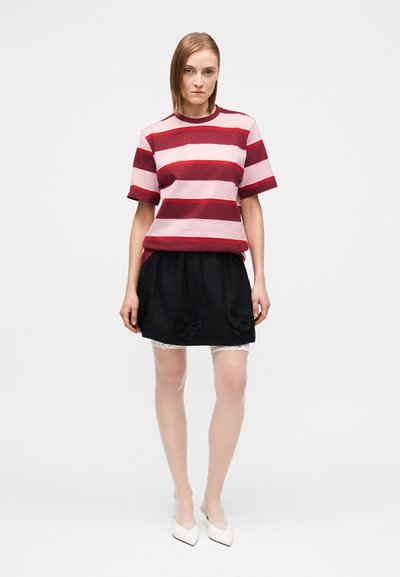 Kobieta ubrana w czerwono-różową top z krótkim rękawem w paski, czarną spódnicę z kwiatowymi detalami, białe rajstopy z koronką i białe szpiczaste buty na obcasie.