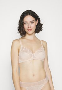 Lichtroze kant beha met zachte cups, bloemenborduursel, verstelbare banden en een delicate strikaccent in het midden. Bijpassende slip zichtbaar.