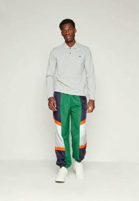 Polo cinza de manga longa com um pequeno logótipo, combinado com calças de treino multicoloridas em verde, azul-marinho, branco e laranja. Sapatos brancos.
