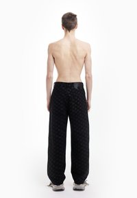 Pantalon large à motif noir avec une finition texturée, comprenant une poche plaquée à l'arrière et des poches lat érales, porté avec des baskets grises.