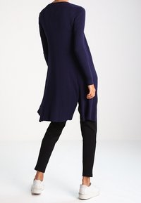 Laivastonsininen ribbineulottu pitkä neule rentolla istuvuudella, jossa pyöreä helma ja pitkät hihat, yhdistetty mustiin housuihin ja valkoisiin tennareihin.