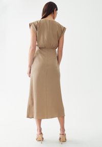 Robe beige, longueur genoux, avec des manches courtes, taille froncée et texture lisse. Fini avec une fermeture éclair au dos, porté avec des sandales à talons tan.