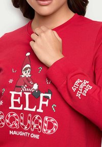 Punainen pitkähihainen paita, jossa on graafinen kuva elfistä, tekstit "ELF SQUAD" ja "Elves Behavin' Badly", karkkikeppikuviot, pehmeää puuvillakangasta.