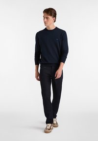 Jersey de cuello redondo azul marino hecho de una tela suave, combinado con pantalones de mezclilla oscuros y zapatillas beiges, con un pequeño logo en el jersey.