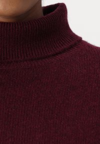 Bordeaux-ribbat stickad polotröja med en mjuk textur och en kroppsnära krage. Mönster är inte synliga.