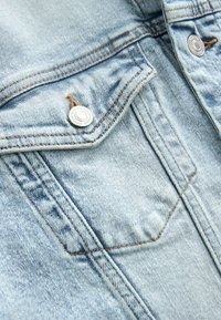 Lichtblauwe denimjasdetail met een borstzak, zilveren drukknoop, zichtbare stiksels en een gestructureerde stofafwerking.