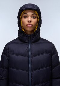 Giacca puffer blu navy con zip frontale e cappuccio foderato in giallo. Presenta cuciture orizzontali e lacci sul cappuccio.
