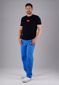 Blaue Baumwollhosen und ein schwarzes T-Shirt mit einem roten Logo. Die Hosen haben ein gerades Bein-Design, und das T-Shirt hat einen klassischen Schnitt. Weiße Sneakers.