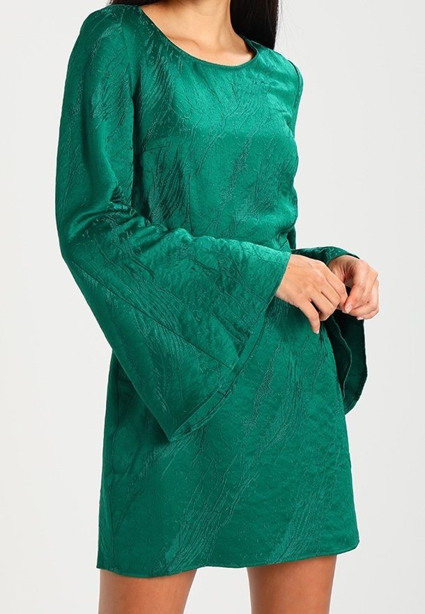 Femme portant une robe verte texturée avec des manches longues évasées, debout les mains croisées devant.