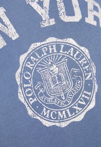 Felpa azzurra con un grafico circolare bianco che presenta una fiamma, un libro e un testo stilizzato che indica "Polo Ralph Lauren" e "MCMLXVII."