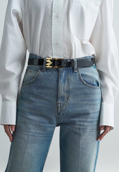 Μαύρη πατεντέ δερμάτινη ζώνη με χρυσό μεταλλικό κράτημα, φορεμένη επάνω από ανοιχτόχρωμα μπλε τζην και ένα άσπρο πουκάμισο με κουμπιά.