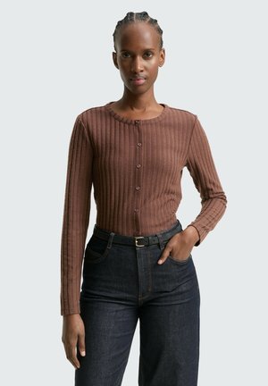 Kobieta z plecionymi włosami, ubrana w brązową prążkowaną bluzkę na guziki z długim rękawem oraz ciemne, wysokie dżinsy z czarnym paskiem, stojąca z ręką w kieszeni.