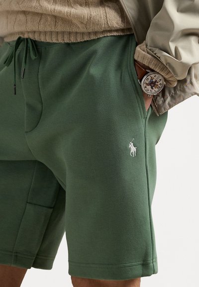 Calções de algodão verdes com cintura elástica e cordão, bolsos laterais e um pequeno logótipo branco. Usados com um pulôver de malha e um relógio.