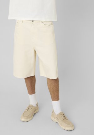 Homme portant un short beige longueur genou, des chaussettes blanches et des chaussures beiges à lacets, debout devant un fond gris clair.
