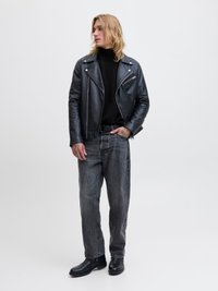 Nuori mies mustassa nahkatakissa, mustassa poolossa, harmaissa farkuissa ja mustissa saappaissa seisomassa, toinen käsi taskussa, katsoo sivulle.