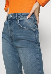 Nahaufnahme einer Person in blauen Jeans und einem orangefarbenen, gerippten Langarmoberteil, wobei eine Hand auf der Hüfte ruht.