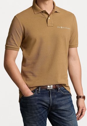 Mand iført en kamelfarvet Polo Ralph Lauren kortærmet poloshirt, blå jeans, brun læderbælte og brunt læderarmbåndsur.