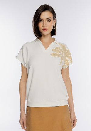 Kobieta o krótkich ciemnych włosach w białym topie z krótkim rękawem ozdobionym beżową haftowaną liściastą aplikacją na ramieniu oraz w beżowej spódnicy.