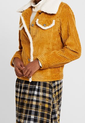 Person iført en sennepsfarvet kordfløjlsjakke med hvidt fåreskindstrim og knapper, kombineret med sorte, hvide og gule ternede bukser.