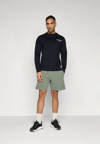 Camisola de gola alta azul-marinho com logótipo, calções verdes, meias brancas e ténis pretos. O conjunto apresenta um corte descontraído e texturas suaves.