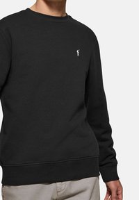 Černá mikina s kulatým výstřihem, žebrovanými manžetami a lemem, s malým bílým logem na hrudi. Vyrobena z měkké, texturované tkaniny.