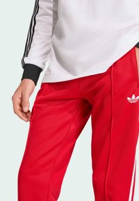 Calças atléticas vermelhas em poliéster com o logotipo da Adidas em branco, riscas laterais e um design de perna reta. Usadas com uma blusa de mangas longas branca.