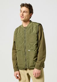 Gilet verde trapuntato con collo rotondo, chiusura a zip, due tasche grandi e un'etichetta con logo. Indossato sopra una maglietta a maniche lunghe color oliva.