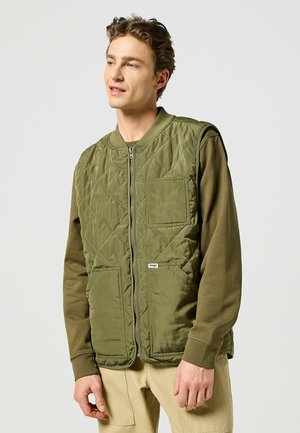 Quilted green vest with a round neck, zip front, two large pockets, and a logo tag. Worn over a long-sleeve olive shirt.