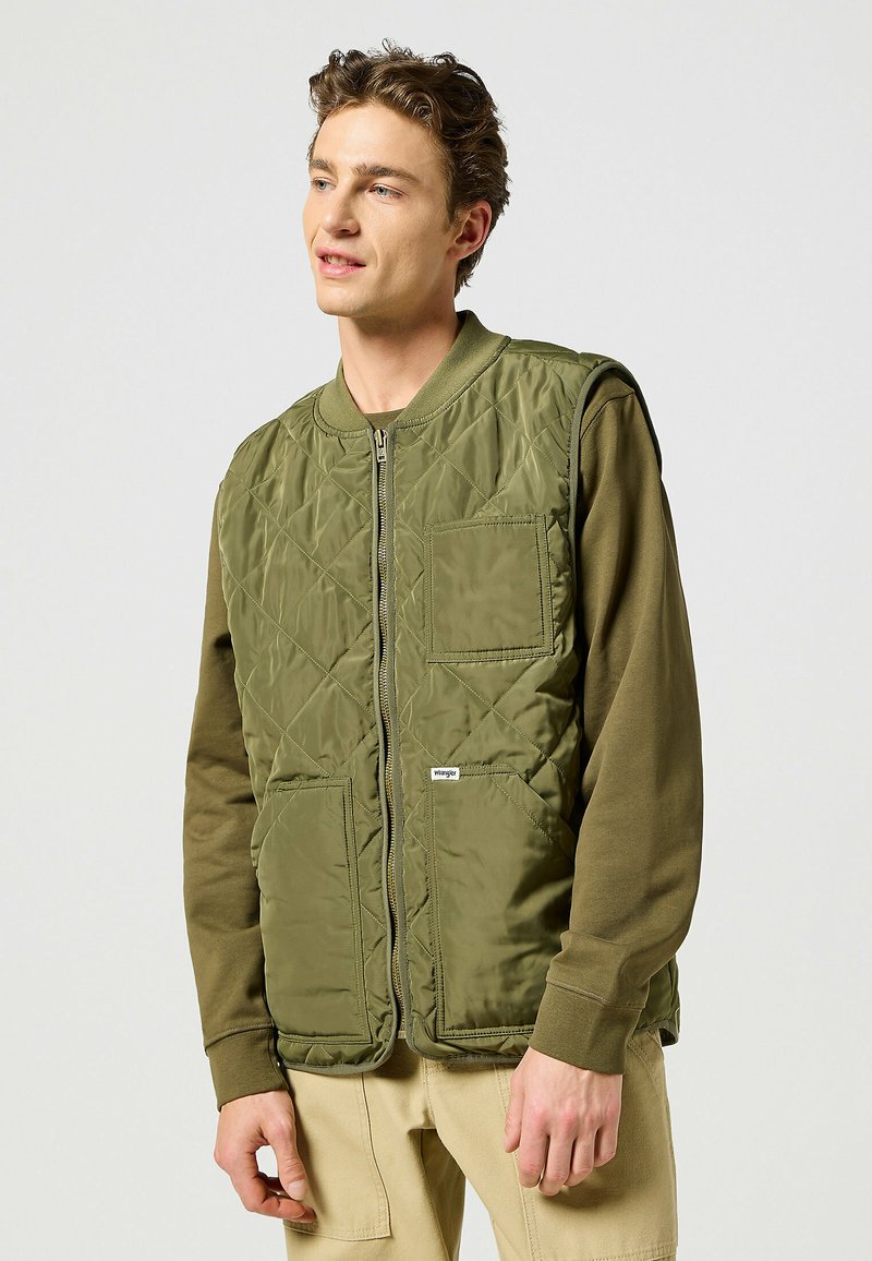 Gilet verde trapuntato con collo rotondo, chiusura a zip, due tasche grandi e un'etichetta con logo. Indossato sopra una maglietta a maniche lunghe color oliva.