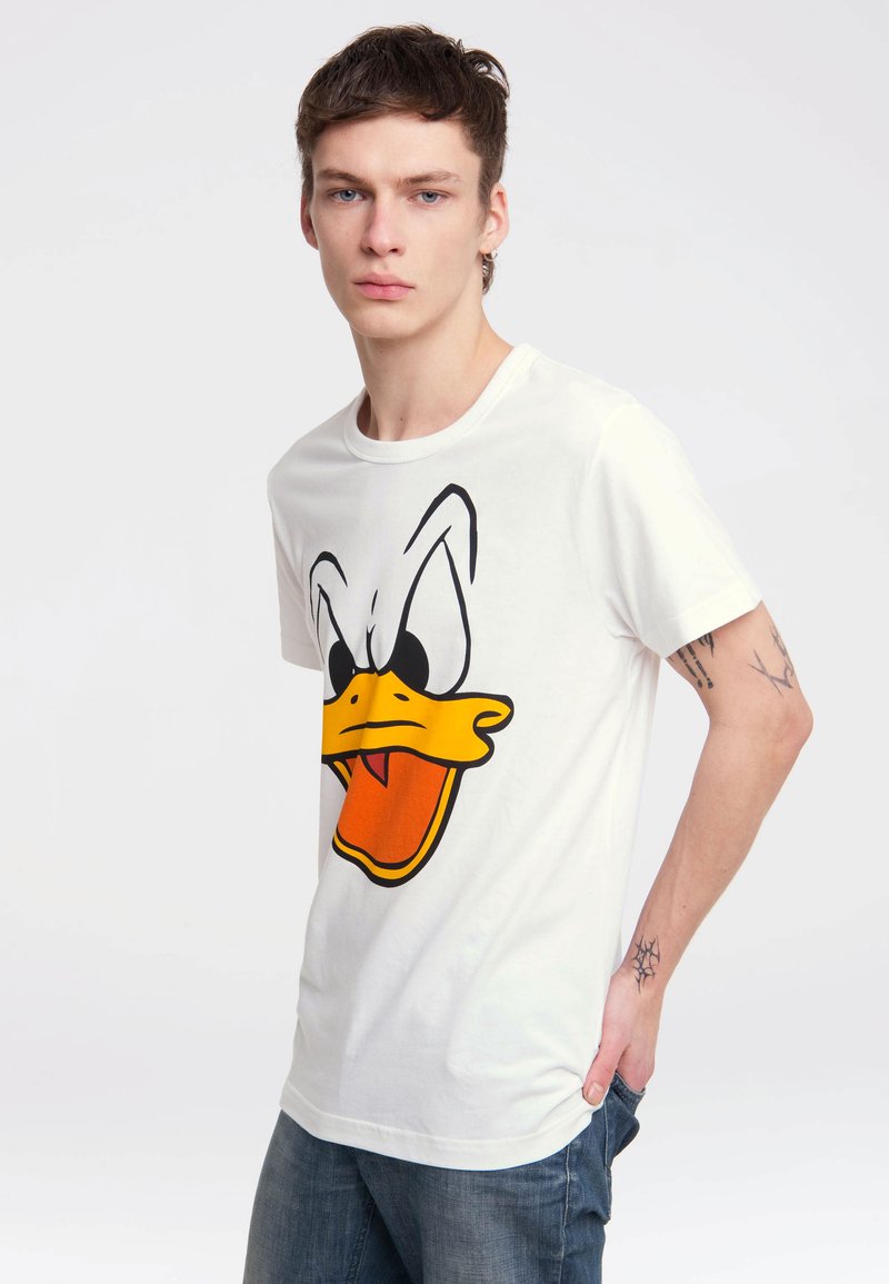 Jeune homme aux cheveux courts portant un T-shirt blanc avec un grand visage de canard de dessin animé et un jean bleu, debout avec une main dans la poche.