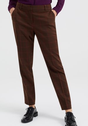 Persoon draagt bruine en rode geruite broek met handen in zakken en zwarte gepolijste veterschoenen tegen een effen achtergrond.