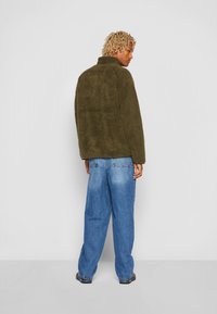 Giacca in pile verde oliva, morbida e pelosa, con colletto alto, spalle abbassate e vestibilità rilassata, abbinata a jeans denim blu larghi.