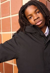 Sort hættejakke med glat tekstur, med lynlås og dekorative trykknapper. Baggrunden er en murstensvæg i jordfarver.