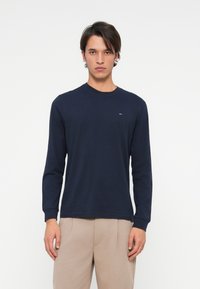 Unge mand iført en navy langærmet skjorte og beige bukser, stående foran en ensfarvet hvid baggrund, vendt mod kameraet med en neutral udtryk.
