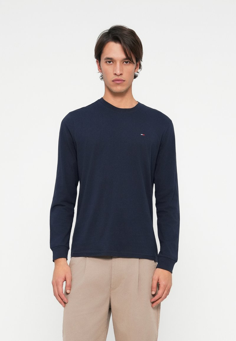 Joven vistiendo una camisa de manga larga azul marino y pantalones beige, de pie contra un fondo blanco liso, mirando hacia adelante con una expresión neutral.