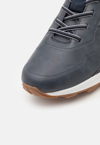 Gros plan sur une basket bleu foncé avec une semelle intermédiaire blanche, une semelle extérieure marron et des lacets assortis bleu foncé sur fond blanc.