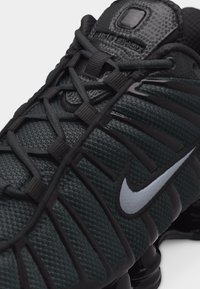 Pantof de alergare Nike negru și verde închis, cu logo alb, partea superioară texturată și șireturi negre care trec prin bucle din material textil.