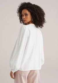 Blouse blanche texturée avec manches bouffantes et encolure ronde, associée à un pantalon rose clair. Mise en avant du tissu et de la coupe du vêtement.