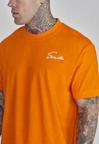 Orange bomulds T-shirt med korte ærmer. Har et hvidt logo på venstre brystområde. Glat tekstur og klassisk rund hals design.